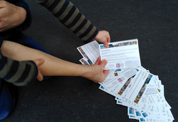 Eine Kinderhand und eine Erwachsenenhand ziehen eine Verlosungskarte aus einem am Boden liegenden Stapel - Copyright: Cornelia Schäfer