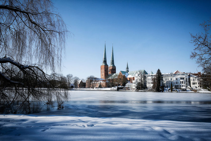 Im Vordergrund der leicht überfrorene Mühlenteich umrandet von Bäumen ohne Blätter. Im Hintergrund der Dom zu Lübeck. Die Türme ragen in den wolkenlosen, blauen Himmel 
