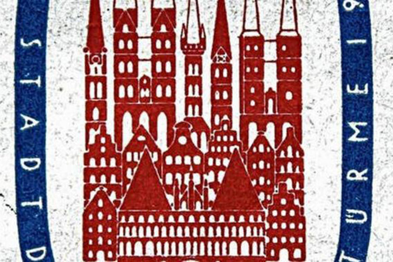 Man sieht den Ausschnitt der Briefmarke von 1956. In der Mitte sieht man in roter Farbe Dächer von Lübeck, sowie die Kirchen und das Holstentor. Umrandet sind diese Häuser von einem blauen breiten Ring, auf dem in weißer Schrift: Stadt und Türme aufgedruckt sind