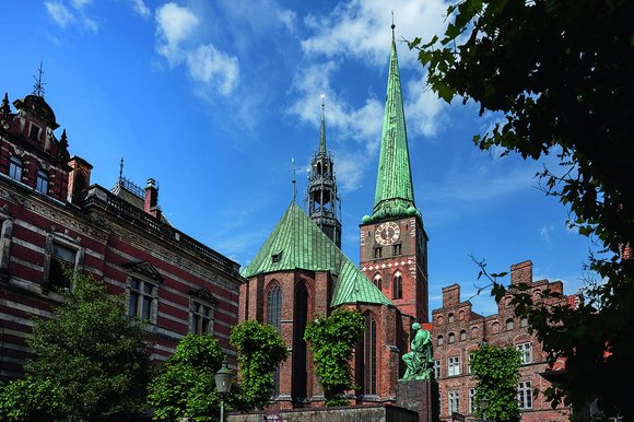 Rechts neben dem Ostchor von St. Jakobi steht das Emanuel Geibel Denkmal | links und rechts von Kirche und Denkmal sind die schönen Altstadtfassaden der Innenstadt zu sehen. Ein paar grüne Bäume sind im Vordergrund zu sehen.
