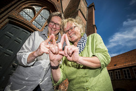 Astrid Lindberg und Heike Schüttler machen das Sieben-Türme-Symbol mit ihren Zeigefingern. Die Finger zeigen eine Turmspitze.