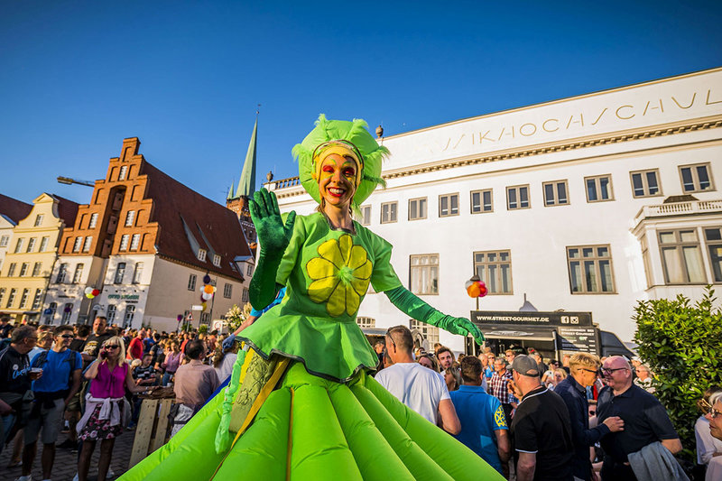 Eine Straßenkünstlerin im grün-gelben Kostum im Vordergrund, hinter ihr eine Menge an Besuchern und die Innenstadtfassaden.