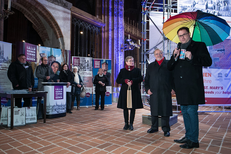 Pröpstin Petra Kallies, Herr Böhning und Bürgermeister Jan Lindenau (mit einem aufgespannten regenbogenfarbenen Regenschirm) stehen nebeneinander in der St.-Marien-Kirche