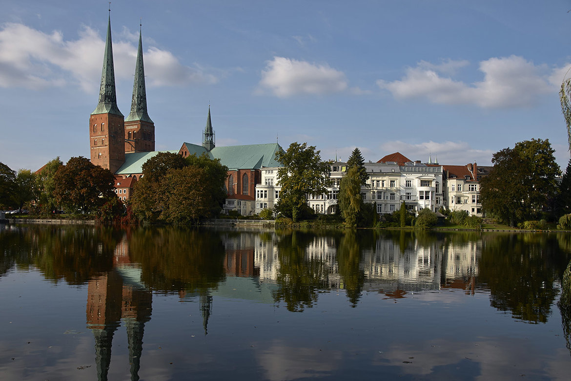 Der Dom zu Lübeck steht neben einer Reihe weißer Häuser. Im Vordergrund ist der Mühlenteich zu sehen, umrandet von grünen Bäumen