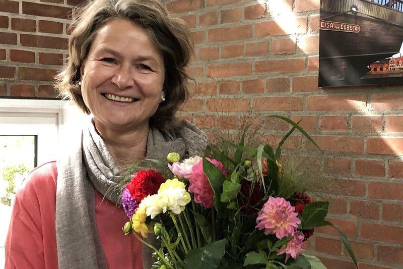 Heike Reimann hält einen Blumenstrauß im Arm und lächelt in die Kamera. Um den Hals trägt sie einen Schal, im Hintergrund sieht man eine Ziegelmauer.
