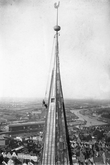 Schwarz-weiß-Fotografie: Zu sehen ist der Handwerker Paul Ruperti, welcher auf einem historischen Hochsitz sitzt und zwar direkt in schwindelnder Höhe am Turmhelm von St. Marien. Das Bild ist von 1929