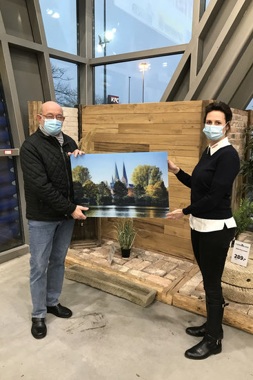 Herr Breede bekommt das Acrylbild überreicht - links Herr Breede, rechts Mitarbeiterin vom Hagebau-Markt Lübeck, zwischen Ihnen halten beide das Bild.