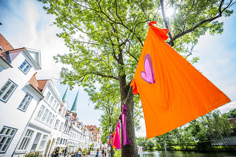 Im Vordergrund ist ein orange-farbenes T-Shirt mit rosa-lila Herz zu sehen, welches auf einer Wäscheleine hängt. Im Hintergrund sieht man einen schönen grünen Baum und die Altstadthäuser der Obertrave, sowie die beiden Turmspitzen des Doms.