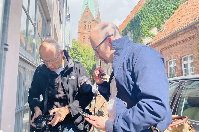 Der Fotograf Rainer Jensen und der Grafiker Markus Endreß schauen auf die Kamera und ins Handy, wie die Bilder geworden sind. Im Hintergrund sieht man einen Kirchturm