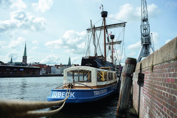 Im Vordergrund ist ein Touristenboot mit der Aufschrift "Lübeck" und "Travemünde" zu sehen, davor liegt ebenfalls im Wasser der Trave das Kraweel, ein mittelalterlicher Schiffstyp aus Holz. Im Hintergrund ragt St. Jakobi über den Dächern der Untertrave, ebenso die Türme von St. Marien