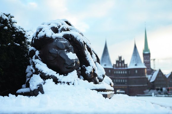 Der Steinlöwe vor dem Holstentor Lübeck ist in einer schlafenden Position. Er ist mit Schnee bedeckt, ebenso das Holstentor im Hintergrund und rechts daneben St. Petri.