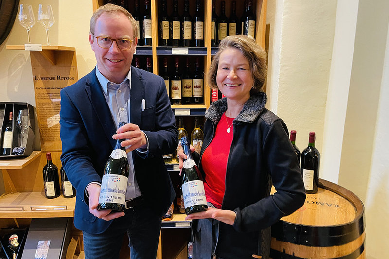 Heike Reimann und Manuel Mack halten jeweils eine Flasche rotwein und eine Flasche Sekt in den Händen. im Hintergrund sind Regale mit Wein- und Sektflaschen und Weingläser zu sehen.