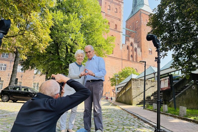 Das Ehepaar Maetzel zeigt mit den Fingern das Sieben-Türme-Symbol, im Vordergrund fotografiert in der Hocke der Fotograf, im Hintergrund ist der Dom zu Lübeck zu sehen