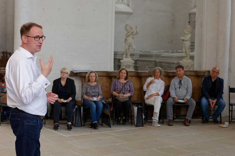 Bürgermeister Jan Lindenau steht im Vordergrund links, ein Teil der Kalenderpaten und Sieben Türme Teammitglieder sitzen auf Holzbänken im Dom zu Lübeck