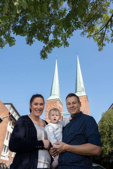 Familie Kosakowski stehen nebeneinander, inder Mitte ihr Kleinkind, im Hintergrund sind die beiden Türme vom Dom zu Lübeck zu sehen. Der Himmel ist strahlend blau und über dem Ehepaar sieht man das grüne Blätterdach eines Baumes