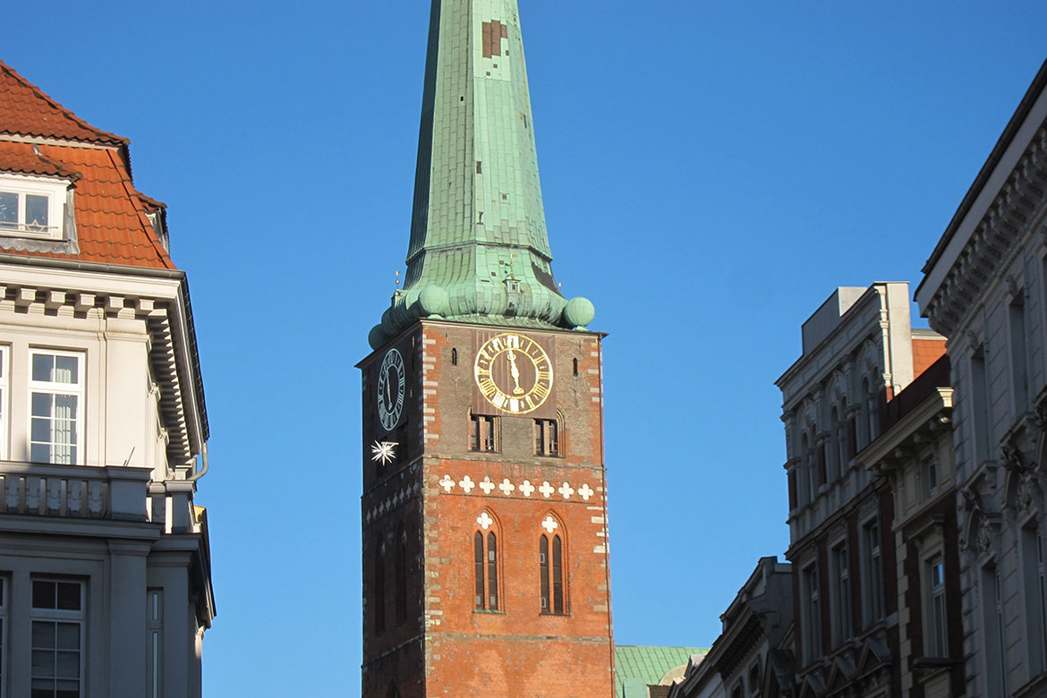 Der Turm von der St.-Jakobi-Kirche ragt in den blauen Himnmel, links und rechts die Fassaden Lübecker Häuser
